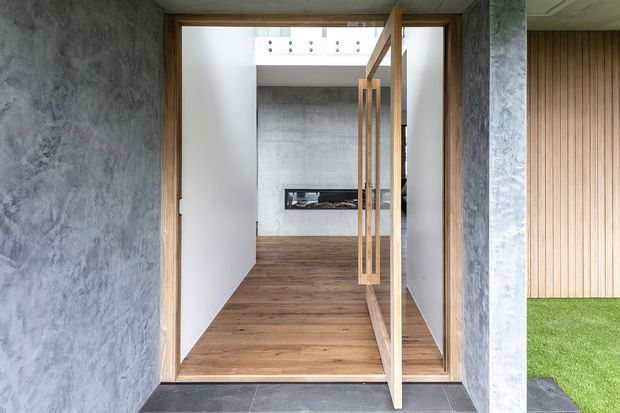 A minimalist residential entrance viewed from outside, featuring a tall timber-framed glass door, textured grey concrete or plaster walls, dark tiled threshold, and a warm timber floor leading into a clean interior. Vertical timber wall panelling and a small grass courtyard are visible to the right, while a recessed dark fireplace or feature opening appears at the rear.
