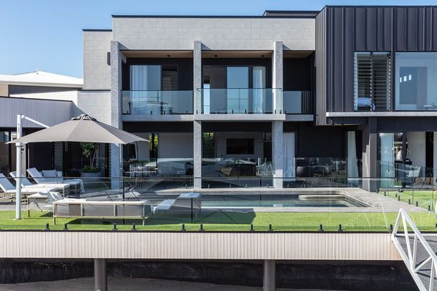 Contemporary multi-storey residential facade overlooking a landscaped terrace and swimming pool. The building combines light grey masonry or concrete-like wall surfaces with dark vertical metal cladding, extensive glazing, glass balcony balustrades, and strong rectilinear volumes. Outdoor seating and an umbrella surround the pool, with a bridge or stair structure visible at the right.