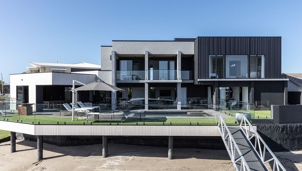 A contemporary multi-storey waterfront residence viewed from the shore, with a dark vertically clad upper volume, concrete-framed balconies, extensive glazing, a large outdoor terrace and swimming pool, and a raised deck connected by a white metal stair and pier structure.