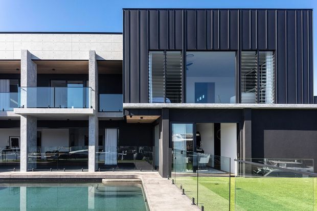 Contemporary multi-storey residential facade with a dark vertically ribbed upper section, exposed concrete structural framing, extensive glazing, glass balcony balustrades, and a ground-level swimming pool with a manicured lawn. The composition emphasizes clean rectilinear forms and strong contrast between charcoal cladding, pale concrete, and blue-green pool water.