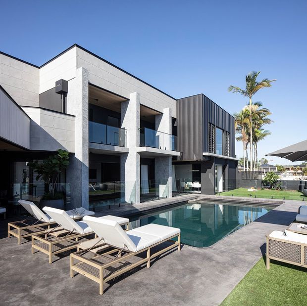 Contemporary multi-level residence with a rectilinear concrete and dark metal facade, large glazed openings, balconies, and an integrated swimming pool courtyard. Palm trees, lawn, outdoor umbrellas, and sun loungers create a resort-like outdoor setting.