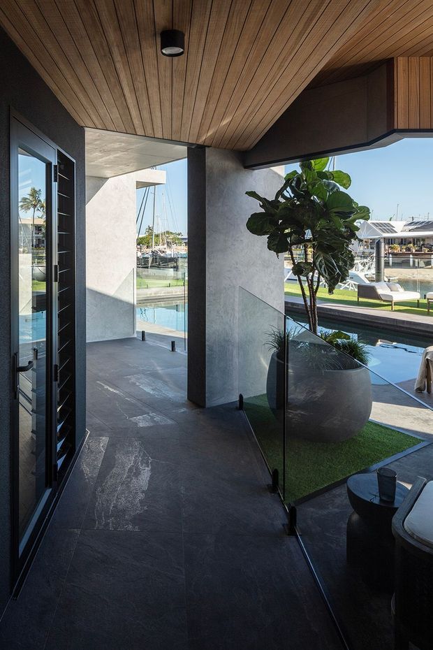 A contemporary covered entry or indoor-outdoor transition space with dark stone flooring, charcoal rendered walls, extensive glazing, and a warm timber-lined ceiling. A glass balustrade encloses a stair or sunken area beside a large potted plant, while the open view reveals a pool and waterfront marina beyond. Black-framed pivot-style glazing and strong geometric forms create a refined, high-contrast architectural setting.