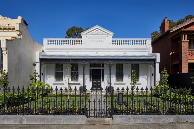 Front elevation of a narrow heritage terrace-style residence with a white rendered facade, ornate parapet and pediment, decorative iron lacework verandah, tall sash windows, and a black wrought-iron front fence and gate. The symmetrical Victorian-era detailing is framed by neighbouring urban homes and a small front garden.