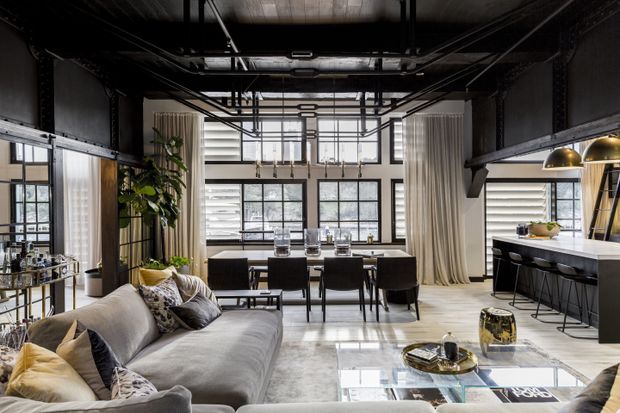Open-plan industrial-style living and dining space with a kitchen at the rear. The room features exposed black ceiling structure and services, steel-framed glazing, dark metal shelving and beams, pale curtains, a large grey sectional sofa, a black dining table with chairs, and a white kitchen island with bar stools. Metallic pendant lights and warm timber accents add contrast to the monochromatic interior.