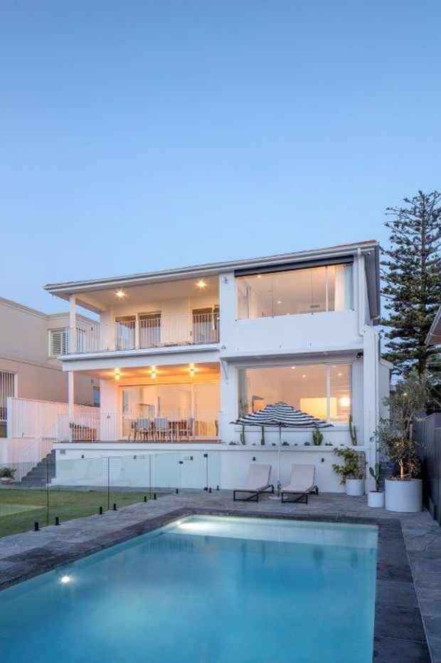 Exterior view of a contemporary multi-storey white residence at dusk, featuring broad balconies, large glazed openings, a paved pool terrace, and a rectangular swimming pool in the foreground. The home has clean horizontal lines, white rendered walls, dark roof edging, outdoor lounge chairs, and a partially visible neighbouring urban residential context.
