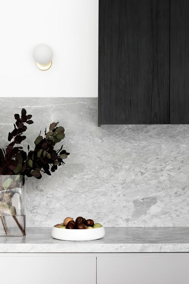 Minimalist kitchen or kitchenette vignette featuring a pale grey-white veined stone benchtop and matching full-height splashback, sleek white lower cabinetry, and dark vertical-grain timber overhead cabinetry. A spherical wall light, decorative vase with foliage, and fruit bowl add restrained decorative accents.