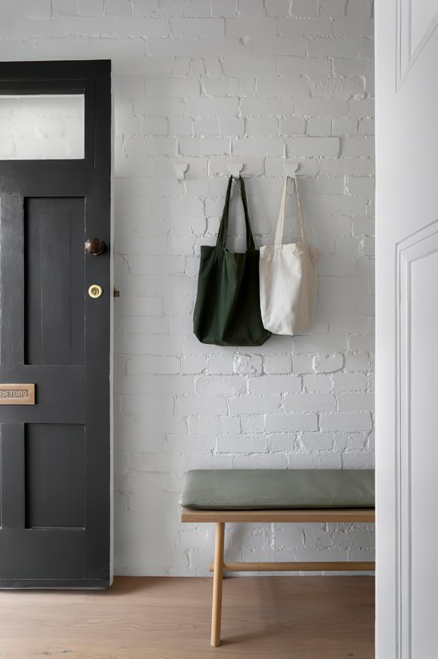 A stylish entryway featuring a black paneled front door, white-painted exposed brick walls, wall hooks with green and cream tote bags, and a simple bench with a muted green upholstered seat and timber legs. Light timber flooring and white trim create a clean, understated interior.