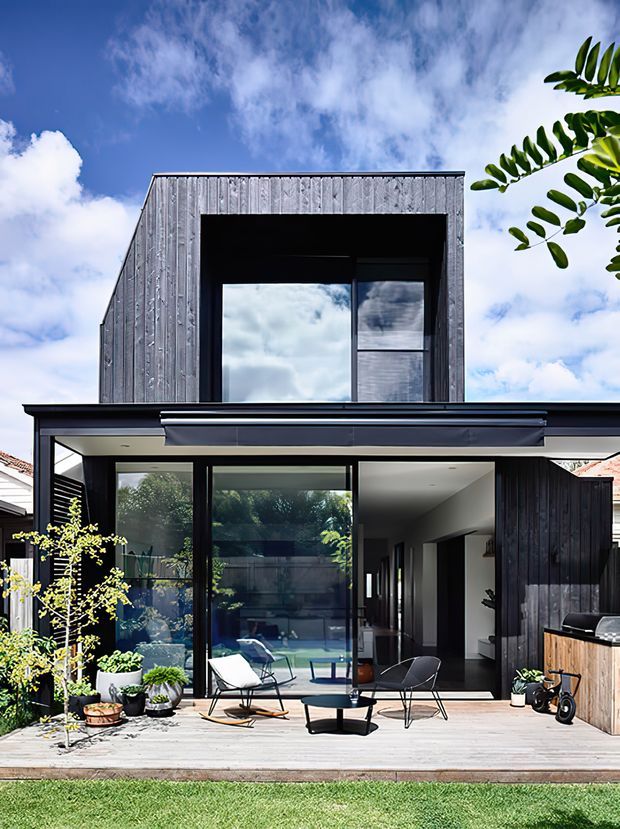 Exterior view of a contemporary two-storey home extension with dark vertical timber cladding, expansive black-framed glazing, and a projecting flat roof over a timber deck. The ground floor opens onto an outdoor sitting area and landscaped courtyard, while the upper level features a large recessed window within a box-like volume.