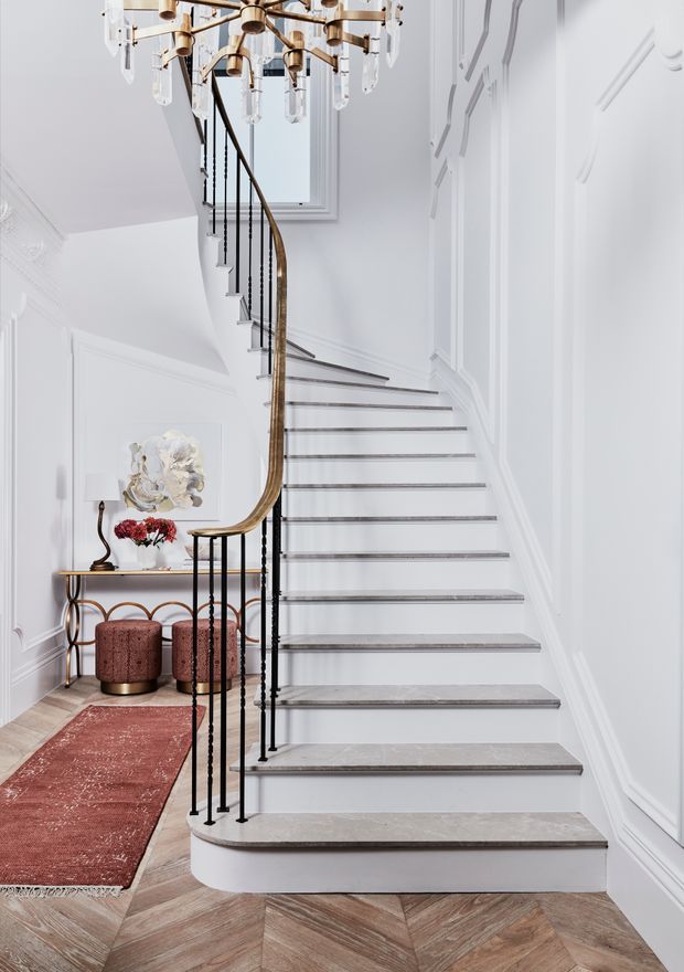 Elegant residential staircase viewed from the entry hall, featuring white decorative wall panelling, a sweeping curved timber handrail with slender black metal balusters, pale stone or concrete stair treads, and a herringbone timber floor. A sculptural brass-and-glass chandelier hangs overhead, with a console table, stools, artwork and rug completing the refined setting.