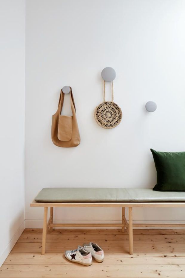 A bright, minimalist entryway with white walls, a light timber floor, and a simple timber bench with a pale upholstered seat. A tan leather bag, woven circular wall hanging, and dark green cushion add natural texture and restrained colour.
