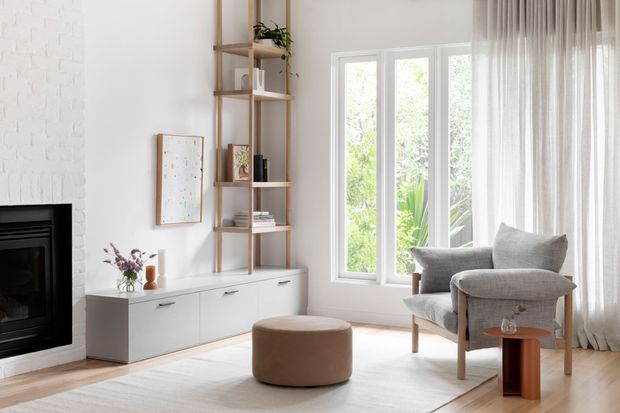 Bright, serene living room with a white-painted brick fireplace, low built-in white media cabinetry, open timber shelving, pale walls, large white-framed windows, sheer curtains, a grey upholstered armchair, and a round tan ottoman. The room has a soft neutral palette and abundant natural light.