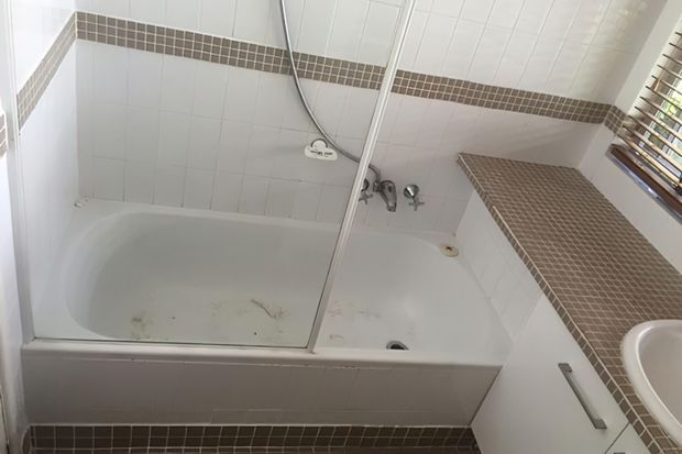 A compact bathroom with a white tiled bath and shower enclosure. Small brown mosaic tile bands accent the white wall tiles, with matching mosaic tiling on the bath surround and floor. A partial white vanity basin, chrome fixtures, and timber venetian blinds are visible.