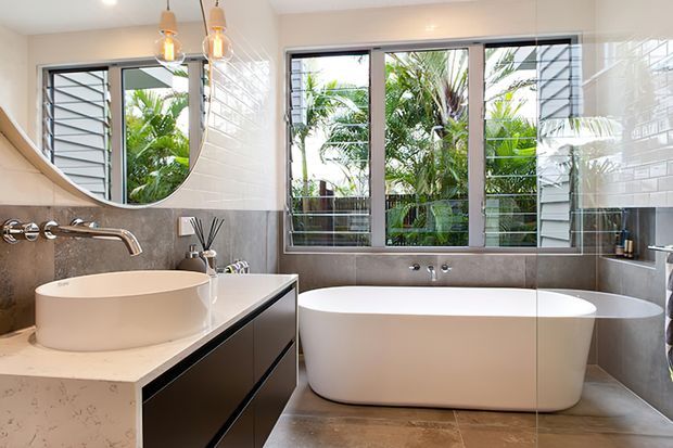 Bright contemporary bathroom with a freestanding white bathtub beneath large louvred windows, a sculptural vessel basin on a stone vanity, glossy white wall tiles, grey tiled lower walls and floor, and pendant lighting. Tropical greenery is visible outside the windows.