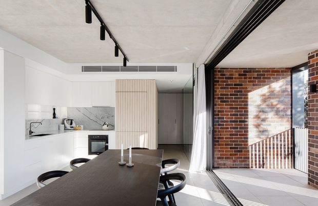 A bright open-plan kitchen and dining area with minimalist white cabinetry, a dark rectangular dining table, and black chairs. A vertically fluted timber-toned pantry or joinery wall forms a feature at the rear, while exposed concrete ceilings, black track lighting, and a large operable glazed opening connect the room to a brick-lined exterior space. Strong daylight creates a clean, contemporary atmosphere.