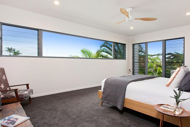 Bright contemporary bedroom with a low-profile timber bed, white bedding, textured grey throw, dark grey carpet, and a vintage upholstered armchair. Wide louvred windows and glazed doors provide abundant natural light and views of tropical vegetation. A white ceiling fan with timber blades and recessed lighting complete the relaxed interior.