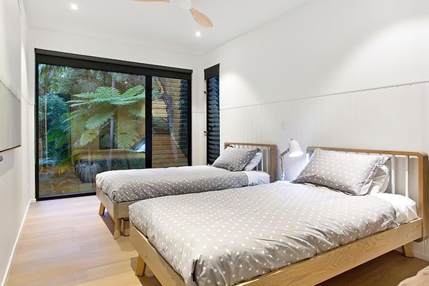 Bright shared bedroom with two timber bed frames, matching grey-and-white polka-dot bedding, bedside lamps, white walls, light timber flooring, and large black-framed glazing with views of lush greenery. A ceiling fan and recessed lighting contribute to the clean, airy feel.