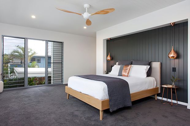 A bright contemporary bedroom with a low timber bed, white bedding, dark charcoal throw, and a dramatic black vertical-panel feature wall. Copper pendant lights hang on either side of the bed, while full-height glazing and louvred windows open to an exterior view. The room has grey carpet, white walls and ceiling, and a timber-bladed ceiling fan.