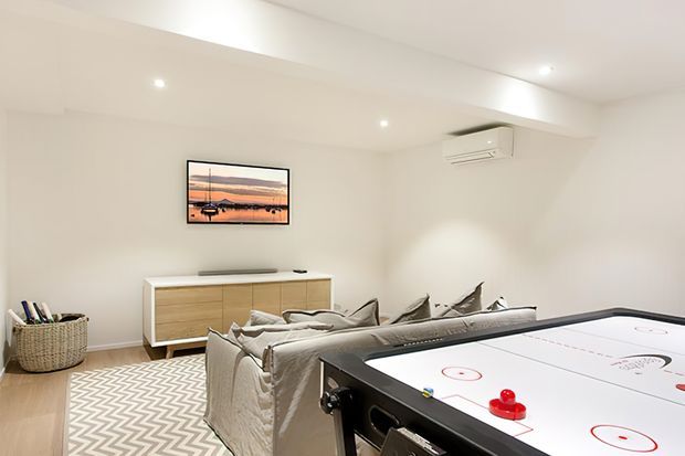Bright contemporary media and recreation room with a large sectional sofa facing a wall-mounted television, a low timber sideboard, and a prominent air-hockey table in the foreground. The room has white walls and ceiling, light timber flooring, a neutral patterned rug, recessed lighting, and minimal decor.