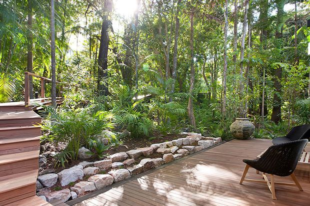 A sunlit timber deck and stairway surrounded by dense tropical woodland, with layered stone garden edging, lush palms and understory planting, and a dark woven outdoor lounge chair. The setting feels secluded, natural, and integrated into the landscape.
