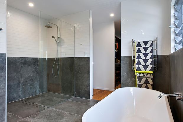 Contemporary bathroom featuring a large frameless glass walk-in shower with dual shower fixtures, dark grey tiled lower walls and floor, white horizontal wall tiles above, and a freestanding white bathtub in the foreground. A heated towel rail with a geometric black-and-white towel sits beside the tub, with timber flooring and a partially visible wardrobe or storage area beyond.