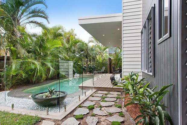 A contemporary tropical residential exterior featuring a narrow swimming pool beside a timber deck and landscaped courtyard. The house has horizontal weatherboard cladding, dark vertical feature cladding, louvred windows, a projecting flat roof and frameless glass pool fencing. Stepping stones, gravel, lush palms and planting create a relaxed outdoor setting.