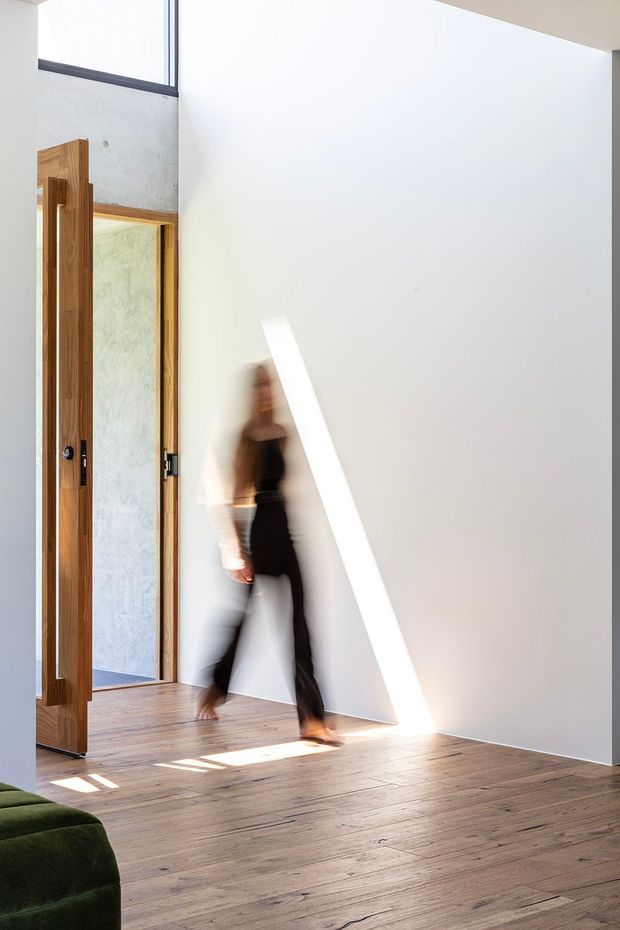 A bright, minimalist interior hallway or entry space with a tall white wall, warm timber flooring, and a large timber-framed pivot door opening toward a concrete-finished room. A strong diagonal beam of sunlight crosses the wall and floor, while a blurred person walks through the space. A small portion of a dark green sofa is visible in the foreground.