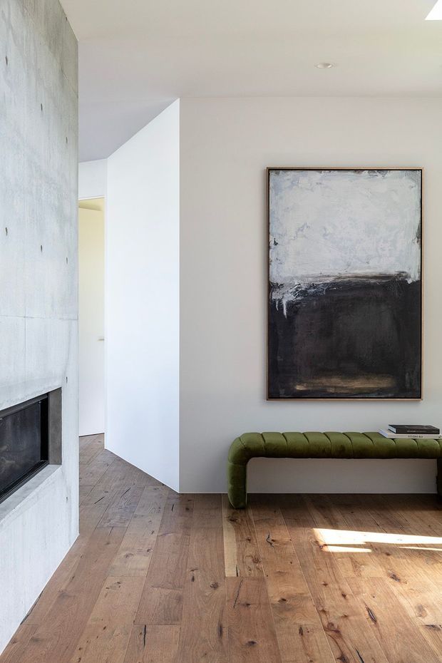 A bright, minimalist interior living area with a white plaster wall, a substantial pale concrete fireplace surround and black fireplace opening on the left, wide-plank timber flooring, and a low olive-green upholstered bench beneath a large abstract artwork. A narrow passage leads deeper into the home, and sunlight falls across the floor.