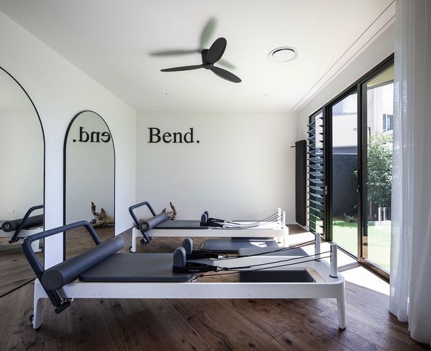 Bright, minimalist home Pilates studio with multiple reformer machines arranged on a warm timber floor. The room has white walls and ceiling, a black ceiling fan, full-height glazed doors with dark frames, sheer curtains, and an arched wall mirror. The word “Bend.” is displayed as wall lettering, and a landscaped courtyard is visible through the glazing.
