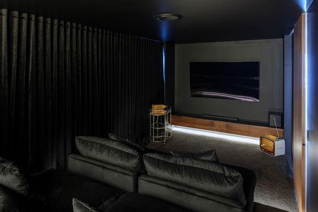 Dark, intimate home cinema or media room with plush sectional seating, black vertical acoustic wall panelling, a large wall-mounted screen, concealed low-level lighting, and a warm timber media console. A small retro-style speaker and decorative shelving add character.