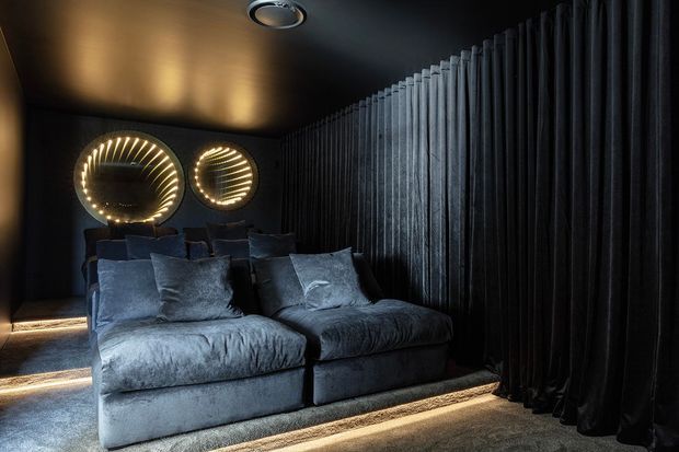 A dark, intimate home cinema or media room with a large plush sectional sofa, dark acoustic-looking vertical wall treatment and curtains, recessed ceiling lighting, and two illuminated circular wall mirrors or feature lights. The room has a luxurious, moody atmosphere with concealed floor-level lighting.