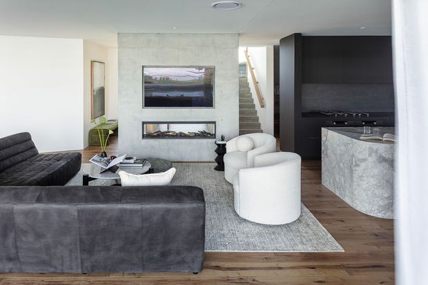 Contemporary open-plan living area with dark leather sofas, rounded white lounge chairs, a textured rug, pale concrete feature wall, and a dark integrated kitchen with a curved stone island. A staircase is visible at the rear, and framed artwork sits above a linear fireplace or dark recess.