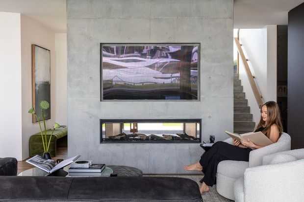 Contemporary living room centred on a grey concrete feature wall with an inset horizontal fireplace and mounted television. A white textured armchair, dark leather ottoman, side tables, artwork, and indoor plant furnish the room, while a stairway and partial kitchen or cabinetry are visible in the background.