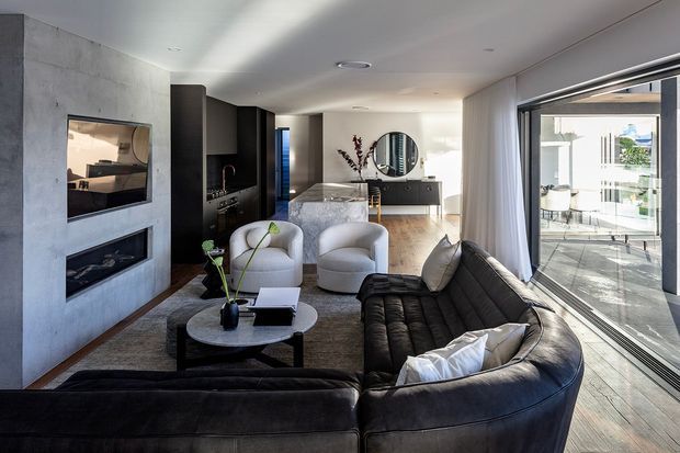 Contemporary open-plan living room with a large black sectional sofa, two light upholstered lounge chairs, and a round coffee table. A concrete feature wall incorporates a television and linear fireplace, while dark built-in cabinetry and a stone kitchen island extend into the background. Full-height sliding glass doors open to a bright outdoor terrace, and timber flooring runs throughout the space.