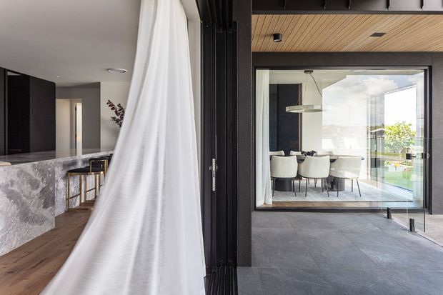 A contemporary residential interior viewed through a partially open black-framed glazed opening. A marble island and dark cabinetry are visible to the left, while a dining area with a light table and upholstered chairs sits beyond the glazing. Large grey floor tiles, timber ceiling lining, white curtains, and strong indoor-outdoor visual connection define the space.