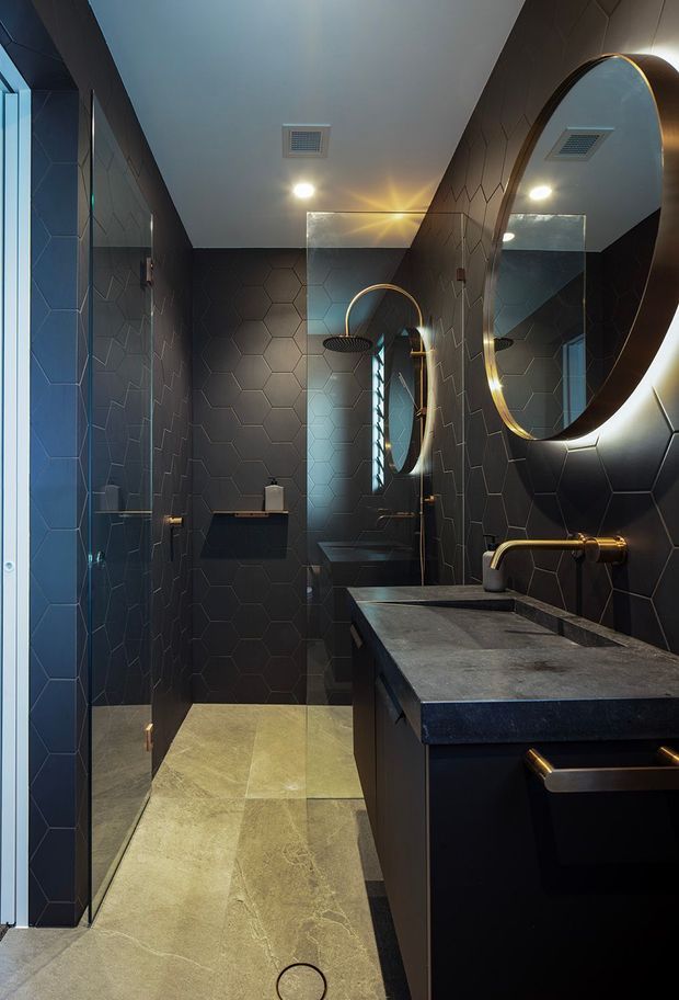 A narrow, dramatic contemporary bathroom with dark geometric hexagonal wall tiles, matching dark cabinetry, a grey stone vanity benchtop, brass tapware and hardware, a large round backlit mirror, and a walk-in glass shower. Pale stone-look floor tiles contrast with the moody walls and joinery.