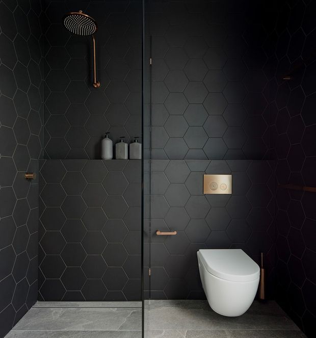 Dark, refined contemporary bathroom with charcoal hexagonal wall tiles, grey stone-look floor tiles, a frameless glass shower enclosure, wall-mounted white toilet, and warm brass/copper-toned shower and hardware. The compact space has a moody, high-contrast atmosphere.