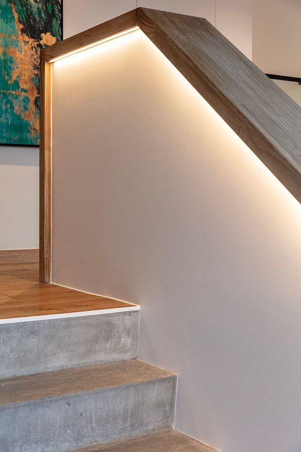 Close-up interior view of a modern stairway with raw concrete steps, warm timber flooring and a timber handrail enclosing a white plasterboard stair wall. Integrated LED strip lighting runs beneath the handrail, creating a warm glow. A partial abstract artwork is visible on the adjacent wall.