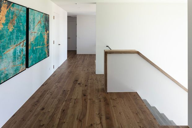 Bright minimalist residential hallway and stair landing with wide warm-toned timber flooring, white walls and built-in doors. A simple timber-trimmed stair balustrade borders the stairwell, while large turquoise and rust abstract artworks add colour along the corridor.