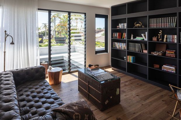 A refined living room with a large tufted dark leather sofa, a distressed dark leather trunk-style coffee table, and extensive floor-to-ceiling black built-in shelving filled with books and decorative objects. Warm timber flooring, neutral walls, sheer curtains, and full-height louvred glazing create a bright, sophisticated interior with views to a planted outdoor area.