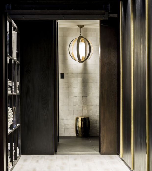 A dark, refined interior passage leads to a compact tiled alcove or entry space. The foreground has pale stone or porcelain flooring, dark timber-toned sliding or panelled doors, open black shelving, and a vertically ribbed metallic wall finish. At the back, pale rectangular wall tiles frame a sculptural spherical pendant light and a glossy dark decorative vessel. The overall composition is moody, luxurious, and highly contrasted, with layered built-in joinery and materials.