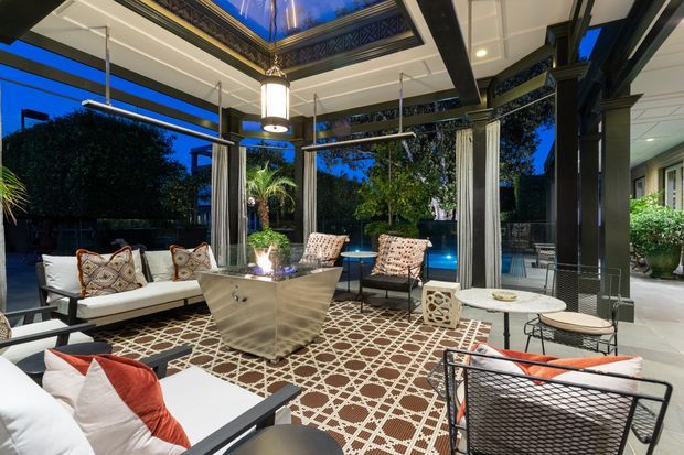A sophisticated covered outdoor lounge at dusk, furnished with sofas, patterned armchairs, a central fire or feature table, and a large geometric rug. The space is enclosed by dark structural framing and decorative metalwork, with a coffered ceiling, pendant lighting, curtains, and views toward a landscaped garden and illuminated swimming pool.