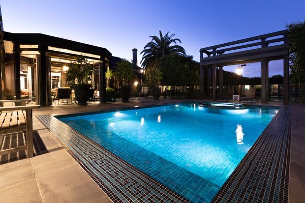 Dusk view of a large illuminated swimming pool surrounded by tiled paving and dark timber decking. A covered outdoor entertaining pavilion with exposed timber framing sits beside the pool, with glazed indoor living spaces visible to the left. Palm trees and planted screening form the background, creating a resort-like outdoor setting.