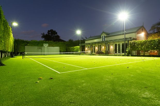 A floodlit outdoor tennis court at dusk, enclosed by tall manicured hedges and mature trees. A timber-clad clubhouse or residential building with a pitched roof, covered veranda, and illuminated windows sits beside the court.