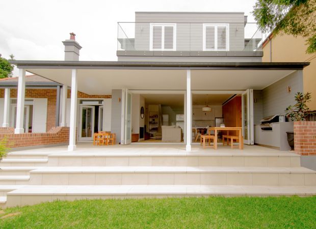 Rear view of a two-storey residential extension with a broad covered terrace and wide-open glazed living areas. The façade combines white-painted detailing, exposed red brick sections, a raised upper level with louvred shutters, and a generous tiled stair terrace leading to the lawn. The open ground floor reveals living and dining areas plus an outdoor kitchen or barbecue zone.
