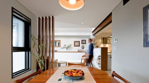 A bright open-plan dining area looking toward a contemporary kitchen and living space. The foreground features a polished warm timber dining table with a fruit bowl, while vertical timber screening separates the dining zone from the adjacent circulation area. A black-framed window, white walls and cabinetry, timber kitchen joinery, exposed timber ceiling beam, recessed lighting and a rounded pendant create a warm, modern interior.