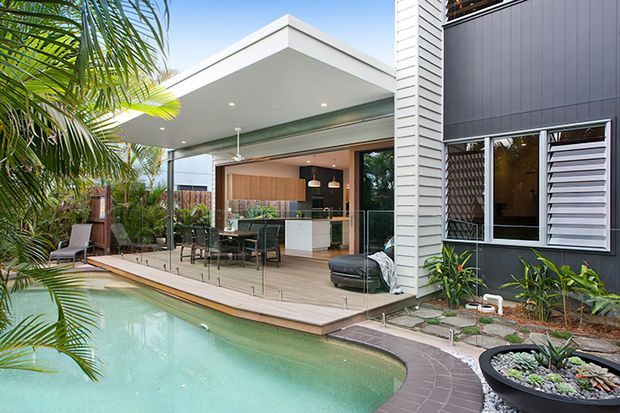 Contemporary tropical residential exterior showing a covered outdoor entertaining deck opening to a kitchen and dining area through extensive glazing. A swimming pool, timber decking, glass balustrade, dark vertical cladding, white weatherboard detailing, and lush palms define the scene.