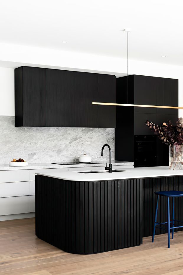 A contemporary kitchen with a large island clad in vertically slatted black cabinetry, white benchtops, and a dark timber-look wall of overhead cabinets. The kitchen includes a pale marble-look splashback, integrated black appliances, a black gooseneck tap, timber flooring, and a slim linear pendant light. A blue bar stool and dried foliage add small accents.