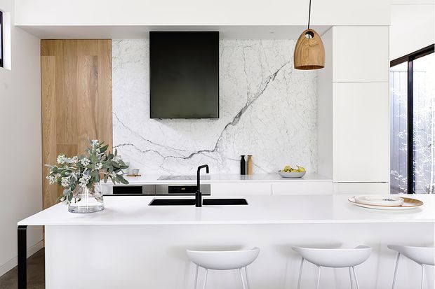 Bright contemporary kitchen featuring a large white island with integrated black sink and tap, white cabinetry, a dramatic white marble-look splashback with grey veining, warm timber pantry cabinetry, and three white bar stools. A black pendant light and black-framed glazing add contrast.