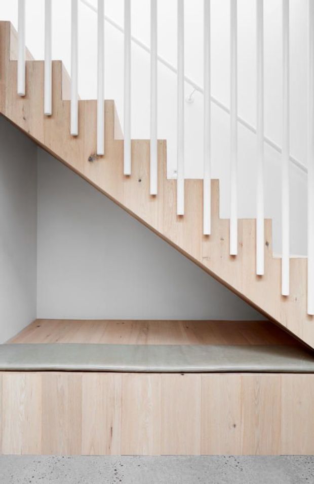 Minimalist interior view beneath a staircase, featuring pale timber stair treads and side panels, slim white vertical balusters, integrated timber cabinetry or panelled storage, a grey upholstered bench-like platform, and a polished concrete floor. The composition emphasizes clean lines, light neutral colours, and efficient use of under-stair space.