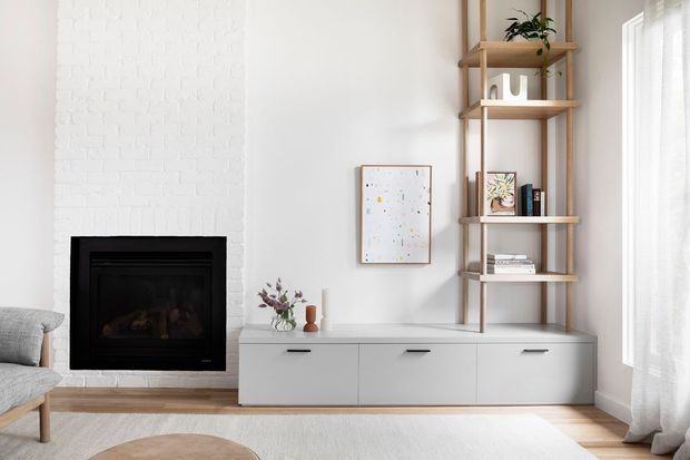 Bright, refined living room featuring a white-painted brick fireplace with black insert, low white built-in cabinetry, an open timber shelving unit, pale timber flooring, and sheer curtains. Neutral furnishings and restrained styling create a clean Scandinavian-influenced look.